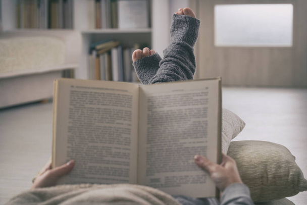 Mujer leyendo un libro con los pies sobre una almohada en casa. Concepto de cuarentena y aislamiento
 - Foto, Imagen