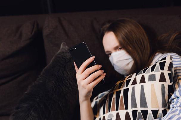 quarantined girl. reading news in a smartphone while lying on a sofa. coronovirus prophylaxis.scientist in biological protective Epidemic virus outbreak concept. COVID-19 - Фото, зображення