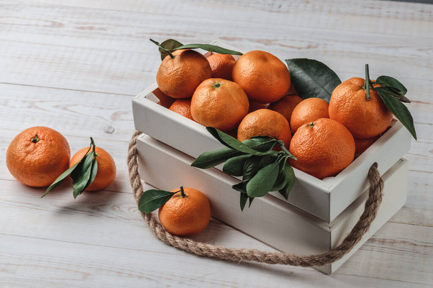 Still-life composition with fresh picked tangerines with green leaves in white wooden box with hemp rope. Textured background with Christmas fruits, side view - 写真・画像