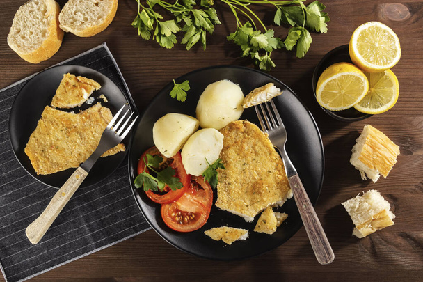 Top view of a plate of delicious baked battered fish seasoned with fresh parsley and lemon and accompanied by cooked potatoes, fresh tomato and freshly baked bread. On a rustic wooden table. - Foto, afbeelding