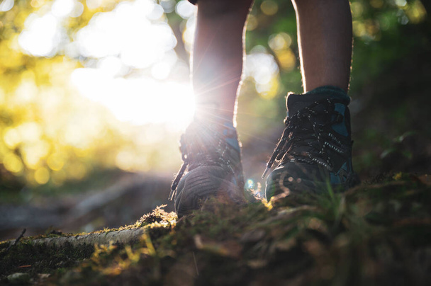 Vista de ángulo bajo de un niño piernas en botas de senderismo de pie en el sendero de montaña iluminado por el sol brillante
. - Foto, imagen