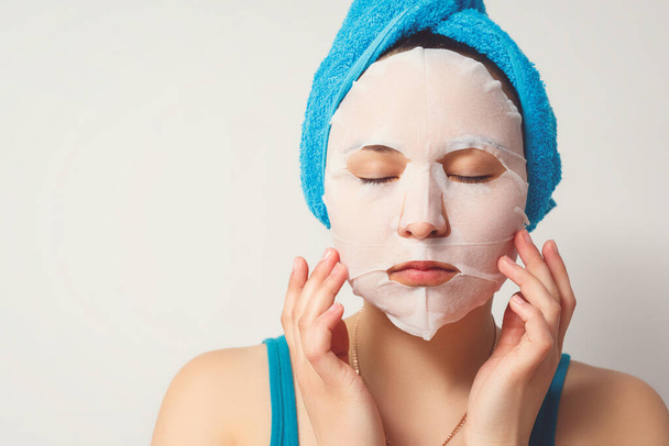 a young beautiful woman uses a moisturizing cosmetic fabric face mask with a towel wrapped around her head. on white background. cosmetic procedures, moisturizing and rejuvenation of the skin. - 写真・画像