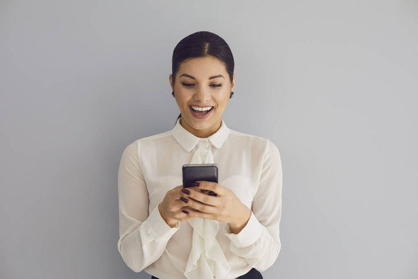 Using the phone on social networks. Hispanic young happy woman typing sms in mobile smiling while standing on gray background. - Φωτογραφία, εικόνα