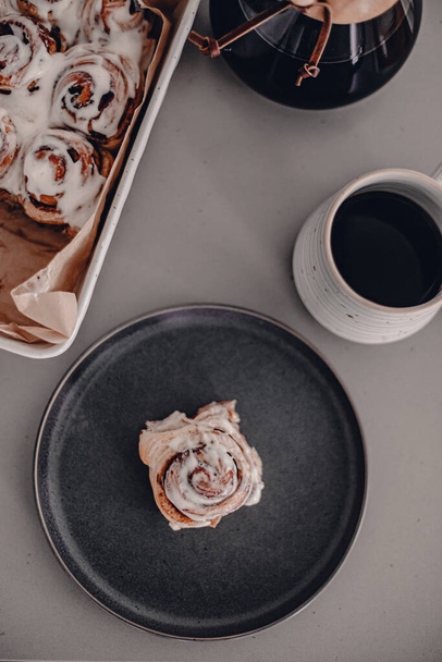 Cinnamon buns with cream cheese frosting. Baked dessert. Cinnamon baked rolls with white glaze. Ceramic baking dish. Breakfast sweet buns with black coffee. Sweet glazed rolls. Grey kitchen table. - Photo, Image