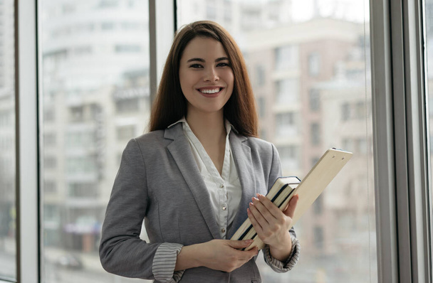 Podnikatelka, držící dokumenty a knihy, stojí v kanceláři u okna. Portrét úspěšného manažera ve formálních šatech při pohledu do kamery a úsměvu - Fotografie, Obrázek