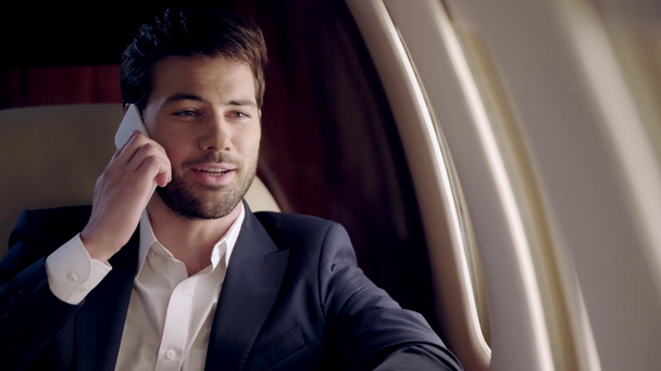 uśmiechnięty, pewny siebie biznesmen rozmawiający na smartfonie podczas podróży samolotem - Materiał filmowy, wideo