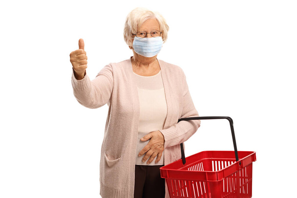 Elderly woman wearing a face medical mask showing a thumb up sign and holding a shopping basket isolated on white background - Fotoğraf, Görsel