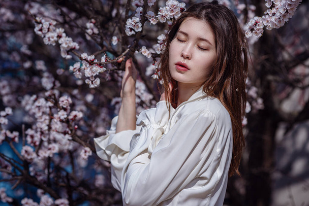 Μια νεαρή Ασιάτισσα σε έναν ανθισμένο κήπο. ρομαντική εικόνα της κομψής casual γυναίκας, μοντέρνα μεταξωτή μπλούζα. θετική διάθεση. Χαρούμενο Ανθισμένο Άνθος στην Ιαπωνία. - Φωτογραφία, εικόνα