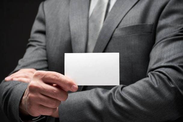 white blank business card closeup in businessman hand, gray suit, dark wall background - Photo, Image