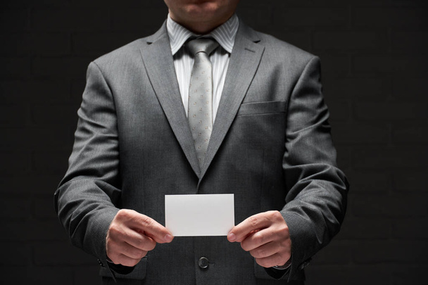 white blank business card closeup in businessman hand, gray suit, dark wall background - Foto, imagen