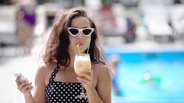 szczęśliwa dziewczyna w okularach przeciwsłonecznych i strój kąpielowy picie koktajlu i za pomocą smartfona - Materiał filmowy, wideo