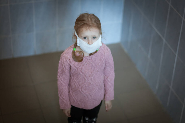 Κορίτσι παιδί είναι λυπημένος, στο πρόσωπό της μια ιατρική μάσκα προστασίας από τον ιό. - Φωτογραφία, εικόνα