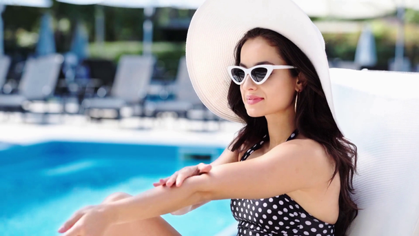 brunette meisje spraying zonnebrandcrème op de hand op ligstoel in de buurt van zwembad - Video