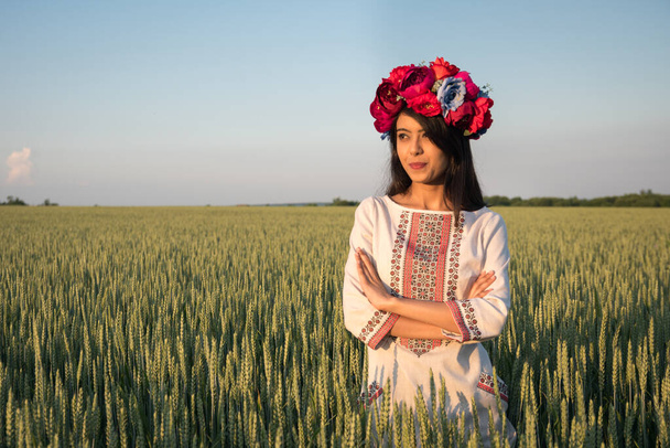 Csinos fiatal nő visel hagyományos ukrán ruhát és virágkoszorú séta a búzamezőn, gyönyörű etnikai lány kézzel készített díszített virágos korona csodálja a természetet, kék ég háttér - Fotó, kép
