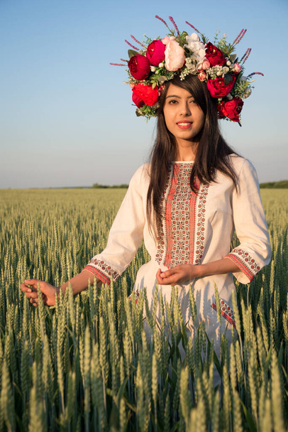 Mooie jonge vrouw dragen traditionele Oekraïense kleding en bloemenkrans lopen in tarweveld, mooie etnische meisje in handgemaakte versierde bloemkroon bewonderen de natuur, blauwe hemel achtergrond - Foto, afbeelding