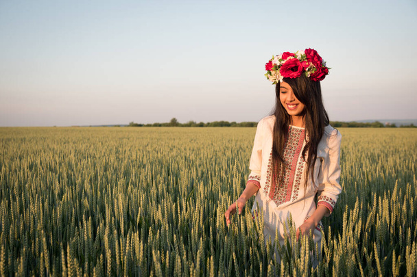 Pretty young woman wear traditional Ukrainian clothes and flower wreath walk in wheat field, beautiful ethnic girl in handmade decorated floral crown admire nature, blue sky background - Photo, Image