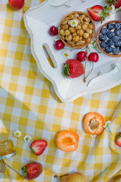 Пикник в парке. Свежие фрукты и пирожные с ягодами и орехами на белом деревянном подносе
 - Фото, изображение