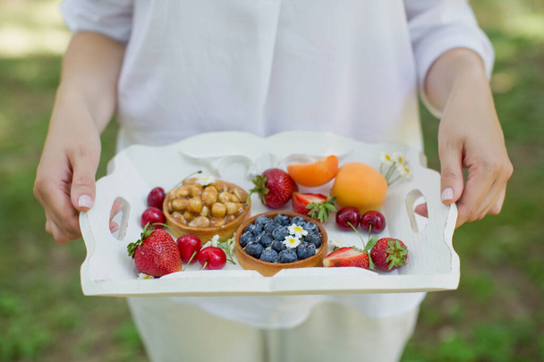 Mujer con ropa blanca sostiene bandeja de madera blanca con frutas frescas y tartas con bayas y frutos secos
. - Foto, Imagen