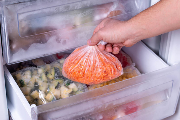 Man putting plastic bag with carrots in refrigerator with frozen vegetables, closeup - Photo, Image