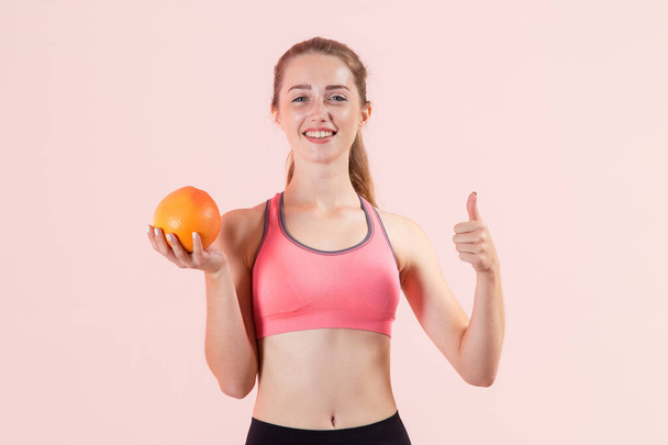 Young sports woman holding a grapefruit in her hands and showing thumb up while standing on a pink background and looking at the camera. Good to eat citrus fruit after a workout. - Foto, Imagem
