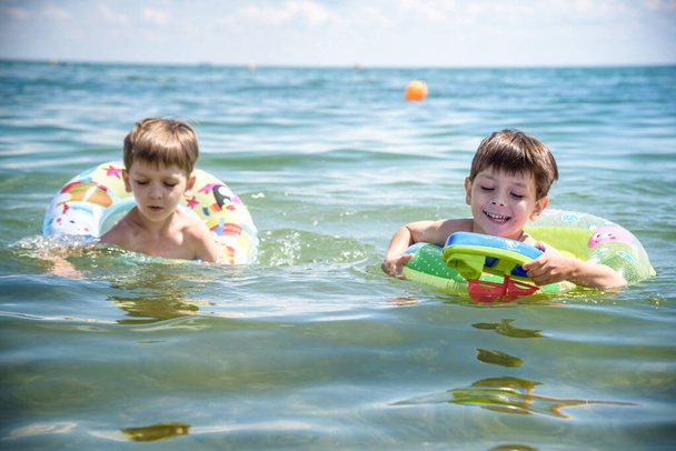 Ευτυχισμένα παιδιά διασκεδάζουν στο θαλάσσιο σερφ στην παραλία. Χαρούμενο ζευγάρι παιδιών σε φουσκωτό δαχτυλίδι βόλτα στο σπάσιμο κύμα. Ταξιδιωτικός τρόπος ζωής, δραστηριότητες κολύμβησης στην οικογενειακή κατασκήνωση. Διακοπές στο τροπικό νησί. - Φωτογραφία, εικόνα
