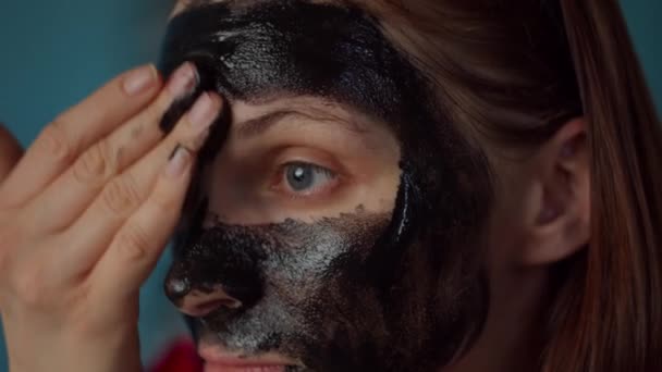 Young 30s woman with black hoop on head applying black cosmetic mask on her face. Woman beauty routine at home on blue kitchen. Close up.  - Materiał filmowy, wideo