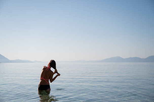 Widok z tyłu młodej kobiety stojącej w spokojnym porannym morzu po kąpieli, ściskającej mokre włosy. - Zdjęcie, obraz