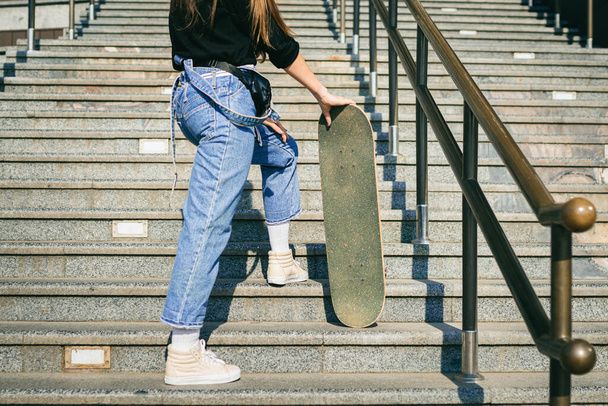 Urban woman with skate. Hipster girl with skateboard in city. Extreme sport and emotions concept. Alternative lifestyle. Stylish hipster girl holding skateboard and posing. Street style outfit. - Photo, Image