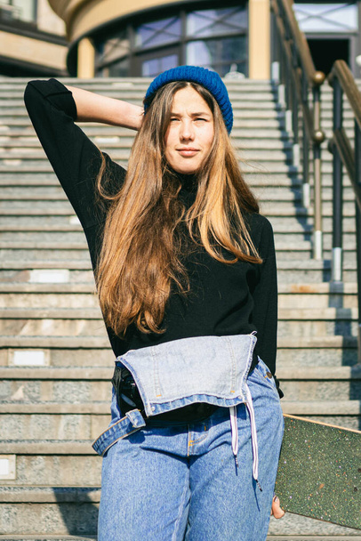 Стильно одягнена жінка в синій джинсовий комбінезон позує зі скейтбордом. Вулична фотографія. Портрет дівчини, що тримає скейтборд. Стиль життя, концепція молоді. Дозвілля, хобі і ковзани в місті
. - Фото, зображення