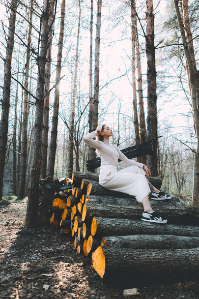 Long haired blonde, standing on the wall of chopped wooden logs, wearing a white linen sweater, white wide pants, headband. Bright sunlight through trees - Fotoğraf, Görsel