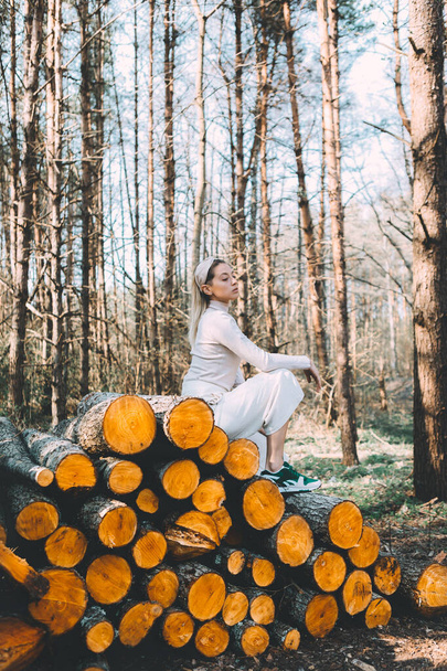 Rubia de pelo largo, de pie en la pared de troncos de madera picados, con un suéter de lino blanco, pantalones anchos blancos, diadema. Luz del sol brillante a través de los árboles
 - Foto, imagen