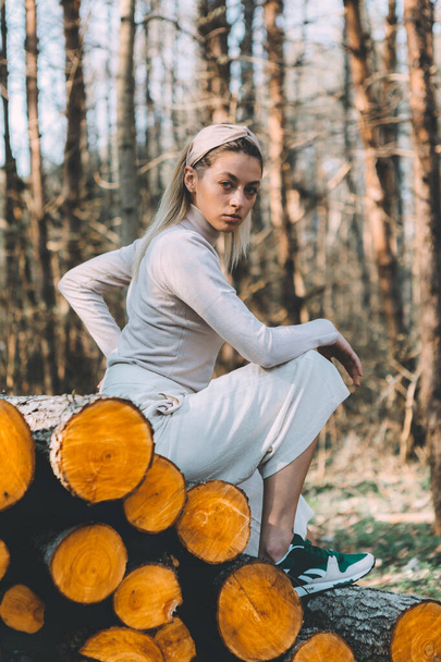 Long haired blonde, standing on the wall of chopped wooden logs, wearing a white linen sweater, white wide pants, headband. Bright sunlight through trees - Fotó, kép