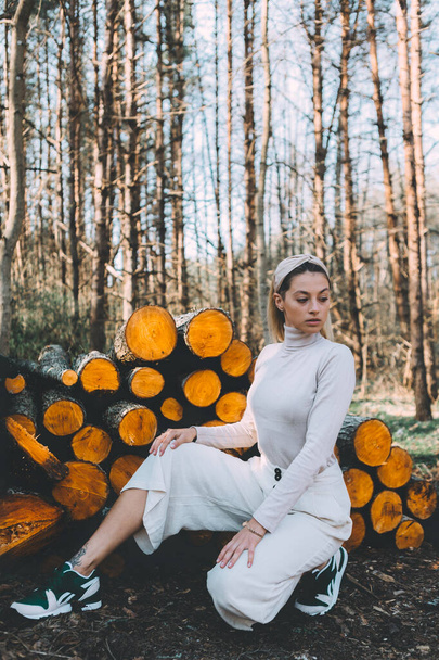 Long haired blonde, standing on the wall of chopped wooden logs, wearing a white linen sweater, white wide pants, headband. Bright sunlight through trees - Photo, Image