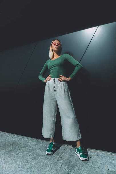 Long haired fashion blonde, standing around the black wall, wearing a green cotton sweater, white wide pants, headband and sunglasses. Bright sunlight flare falls on background - Foto, Imagem