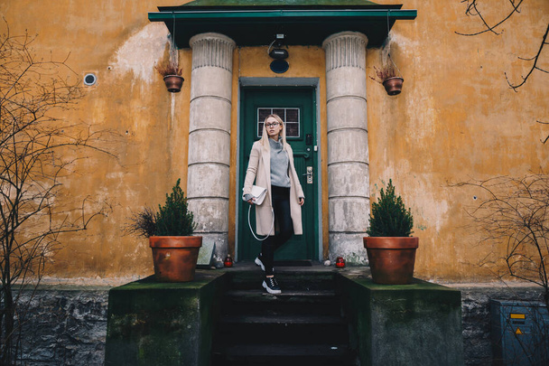 Mulher loira de cabelos compridos usando um casaco de areia com suéter de lã e jeans pretos, em pé em uma escada atrás da casa à moda antiga. Local místico
. - Foto, Imagem