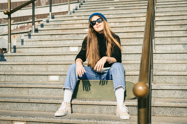 Jeune femme caucasienne posant dans la rue avec planche à roulettes dans les mains. Adolescente en jeans bleus sports extrêmes dans un environnement urbain. Thème des loisirs des jeunes, mode de vie. Skateboarder dans la ville
. - Photo, image