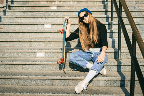 スケートボードとポーズをとるブルーデニムジャンプスーツのスタイリッシュな服を着た女性。ストリートフォト。スケートボードを保持する少女の肖像画。ライフスタイル、若者のコンセプト。市内のレジャー、趣味、スケート. - 写真・画像