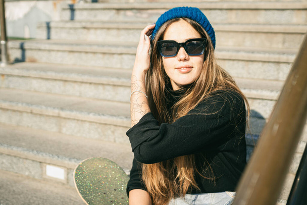 Jonge blanke vrouw poseert op straat met skateboard in handen. Tiener meisje in blauwe jeans extreme sporten in een stedelijke omgeving. Thema van jeugdrecreatie, levensstijl. Skateboarder in de stad. - Foto, afbeelding