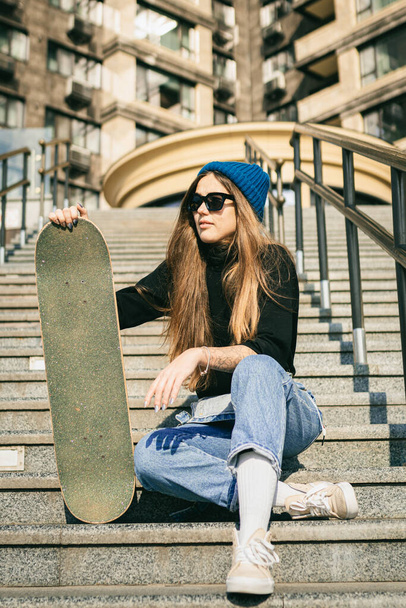 Stylishly dressed woman in blue denim jumpsuit posing with skateboard. Street photo. Portrait of girl holding skateboard. Lifestyle, youth concept. Leisure, hobby and skate in the city. - Foto, Imagem