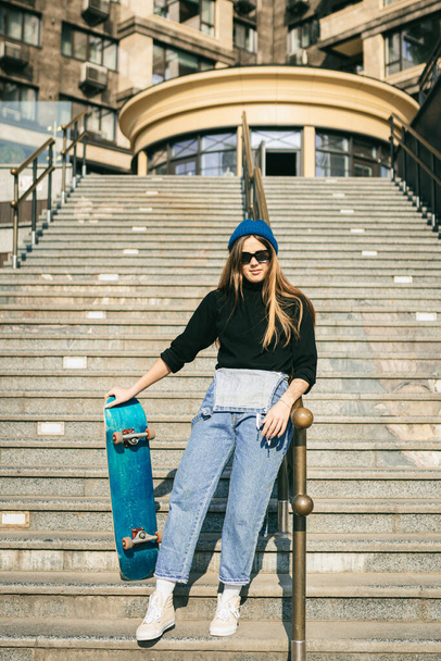 Νεαρή λευκή γυναίκα ποζάρει στο δρόμο με σκέιτμπορντ στα χέρια. Έφηβη κοπέλα με μπλε τζιν extreme sports σε αστικό περιβάλλον. Θέμα αναψυχής των νέων, τρόπος ζωής. Skateboarder στην πόλη. - Φωτογραφία, εικόνα
