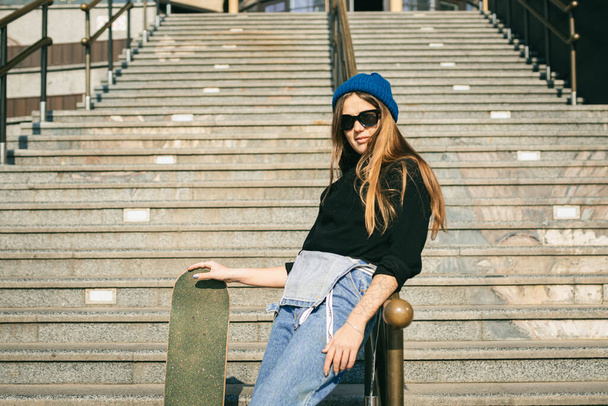 Молода біла жінка позує на вулиці зі скейтбордом в руках. Дівчина-підліток в блакитних джинсах екстремальні види спорту в міському середовищі. Тема молодіжного відпочинку, способу життя. Скейтбордист у місті
. - Фото, зображення