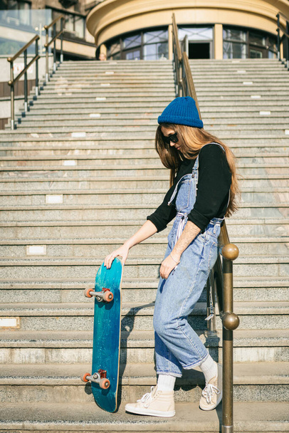 Femme élégamment habillée en combinaison en denim bleu posant avec planche à roulettes. Photo de rue. Portrait de fille tenant skateboard. Mode de vie, concept jeunesse. Loisirs, passe-temps et skate en ville
. - Photo, image