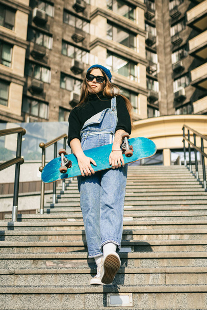 Urban woman with skate. Hipster girl with skateboard in city. Extreme sport and emotions concept. Alternative lifestyle. Stylish hipster girl holding skateboard and posing. Street style outfit. - Foto, Imagem