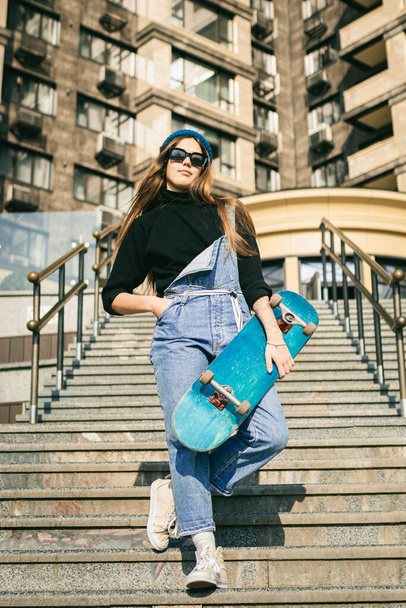 Stylishly dressed woman in blue denim jumpsuit posing with skateboard. Street photo. Portrait of girl holding skateboard. Lifestyle, youth concept. Leisure, hobby and skate in the city. - Фото, зображення