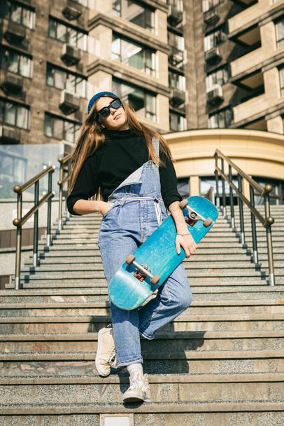 Mladá běloška pózuje na ulici se skateboardem v ruce. Teenager dívka v modrých džínách extrémní sporty v městském prostředí. Téma rekreace mládeže, životní styl. Skateboardista ve městě. - Fotografie, Obrázek