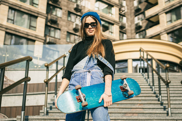 Stylishly dressed woman in blue denim jumpsuit posing with skateboard. Street photo. Portrait of girl holding skateboard. Lifestyle, youth concept. Leisure, hobby and skate in the city. - Foto, afbeelding