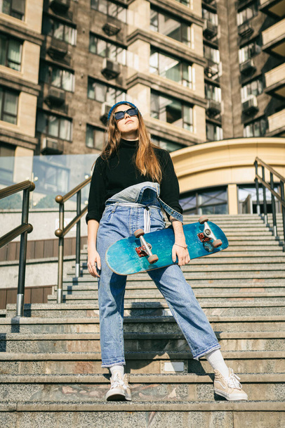 Κομψά ντυμένη γυναίκα σε μπλε denim jumpsuit ποζάρουν με skateboard. Φωτογραφία δρόμου. Πορτρέτο του κοριτσιού που κρατά skateboard. Τρόπος ζωής, έννοια της νεολαίας. Αναψυχή, χόμπι και πατινάζ στην πόλη. - Φωτογραφία, εικόνα