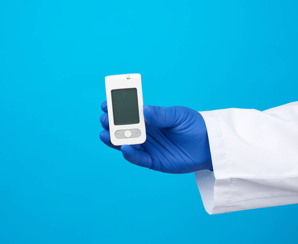 male hand with blue sterile gloves holds a white blood glucose meter for measuring blood sugar, blue background - Photo, Image