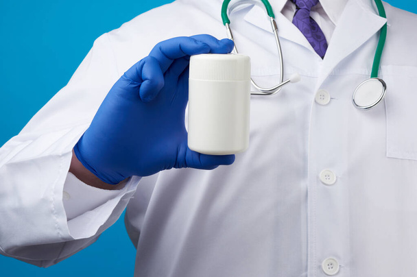 männliche Hand in blauen sterilen Handschuhen hält ein weißes Plastikgefäß für Pillen, medizinisches Behandlungskonzept für Krankheiten, blauer Hintergrund - Foto, Bild