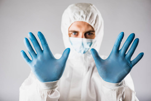 a virologist in a protective suit with a respirator face mask and blue gloves with fear on his face shows cautionary hands with his hands to the camera. collapse of the healthcare system due to the epidemic coronavirus. - 写真・画像